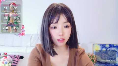 Media: Video of a young East Asian woman with straight black hair, light skin, and a soft expression, wearing a brown sweater, in a bright, minimalist room with white walls, pink flowers, and a Starry Night painting.