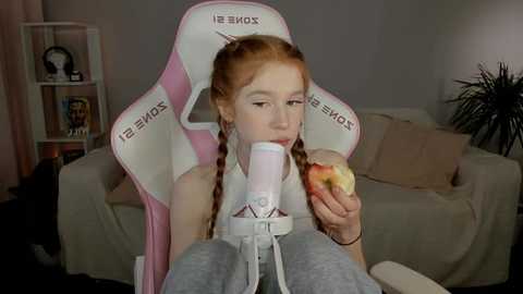 Media: A video of a young girl with red braids, wearing a white \"XBOX\" chair, eating a sandwich in a cozy living room with a beige couch and potted plant.