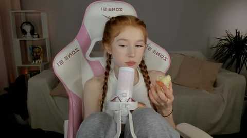 Media: Video of a young girl with fair skin and red braided hair, wearing a white tank top and gray sweatpants, sitting in a pink gaming chair, eating a banana.