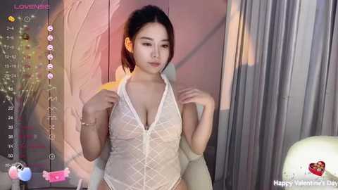 Media: Video of an East Asian woman with dark hair in a ponytail, wearing a sheer white lace bodysuit, standing in a dimly lit room with pink and gray curtains.