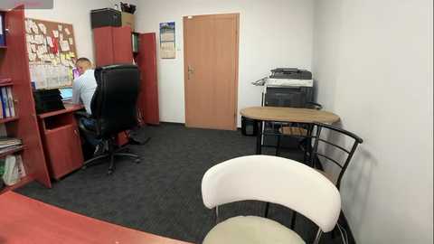 Media: Video of a modern office with a beige chair, wooden desk, and red bookshelves. A man in a white shirt sits at a black desk, facing a computer monitor.