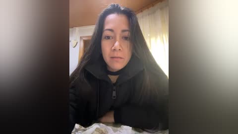 Media: Video of a young woman with long, dark hair, wearing a black hoodie, standing in a dimly lit room with beige walls and white curtains.