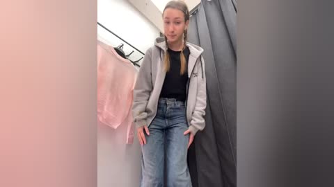 Media: Video of a young Caucasian woman with long blonde hair, wearing a gray hoodie, black shirt, and high-waisted jeans, standing in a changing room with pink and gray clothes hanging nearby.