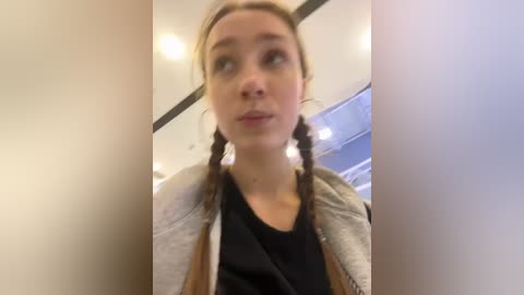 Media: A video of a young woman with fair skin and light brown hair in braids, wearing a grey jacket over a black shirt, looking slightly off-camera in an indoor setting.