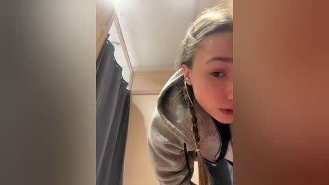 Media: Video of a young woman with fair skin, brown hair, and a grey jacket, peering around a corner into a dimly lit hallway with beige walls and a dark curtain.