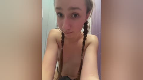 Media: A video of a topless young woman with fair skin and brown hair in braids, taken from a close-up angle, showing her face and upper body.