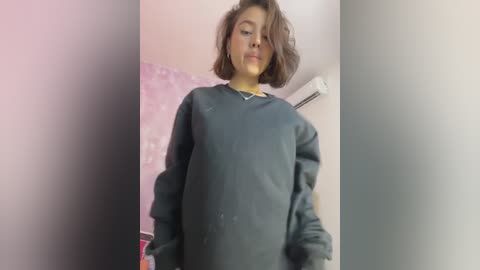 Media: Video of a young person with shoulder-length, wavy brown hair, wearing a dark grey long-sleeve shirt, standing in a room with pink and white walls, and a hanging air conditioner.