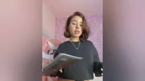 Media: Video of a young woman with light brown hair, wearing a black long-sleeve shirt, standing in a pink room with a white bed and patterned wallpaper. She holds a tablet in front of her.