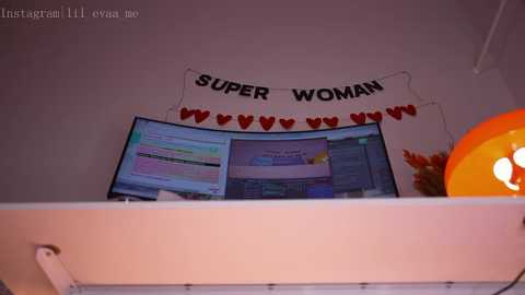 Media: Video of a modern office desk with a white background, featuring a monitor displaying an internet page, a banner reading \"SUPER WOMAN,\" and an orange lamp.