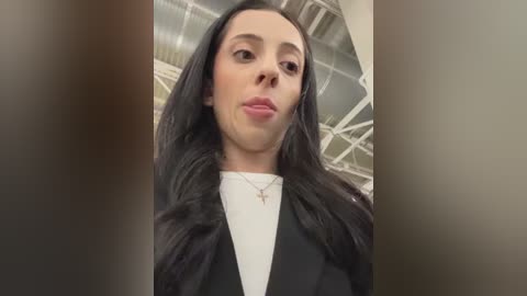 Media: Video of a young woman with long, dark hair, wearing a black blazer over a white t-shirt, standing in a modern, industrial setting with metallic structures in the background.