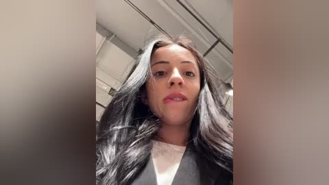 Media: Video of a young woman with long, dark, wavy hair, light skin, and full lips, wearing a black jacket over a white top, taken indoors with blurred background.