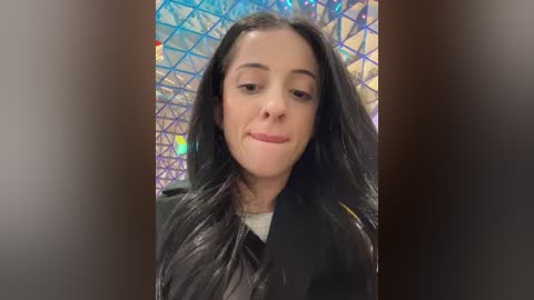 Media: Video of a young woman with long black hair, wearing a black jacket, smiling slightly, in a modern indoor setting with a geometric, multi-colored background.