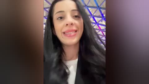 Media: Video of a smiling young woman with long, dark hair, wearing a black blazer over a white shirt. Background features a geometric, reflective pattern in purple and blue hues.