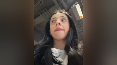 Media: Video of a young woman with long, wavy black hair, wearing a white t-shirt, standing in a dimly lit, industrial room with exposed pipes and a fluorescent light fixture.