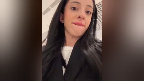 Media: Video of a smiling young woman with long black hair, wearing a black blazer over a white shirt, in a modern, bright room with vertical blinds.