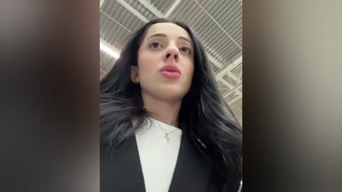 Media: Video of a young woman with long black hair, wearing a black blazer over a white shirt, standing in a modern, industrial building with metal ceilings and bright lights.