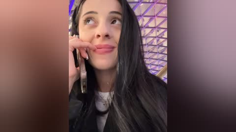 Media: Video of a woman with long, straight black hair, wearing a black jacket, talking on a smartphone, with a geometric purple and white patterned wall in the background.