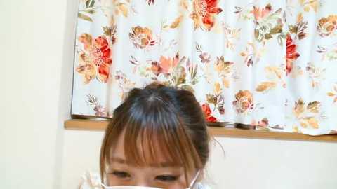 Media: Video of a young Asian woman with light brown hair styled in a bun, wearing a white floral-patterned headband. She is in a room with a white curtain featuring large, colorful flowers.