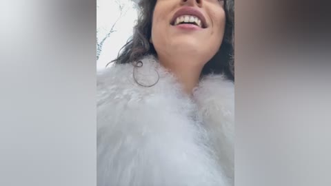 Media: Video of a woman with medium skin tone, wearing a fluffy white coat and black-framed glasses, smiling broadly. Background shows blurry, out-of-focus trees.