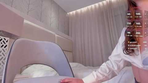 Media: Video of a woman in a white shirt lying on a bed with light gray walls and a headboard, in a modern, minimalist bedroom setting.