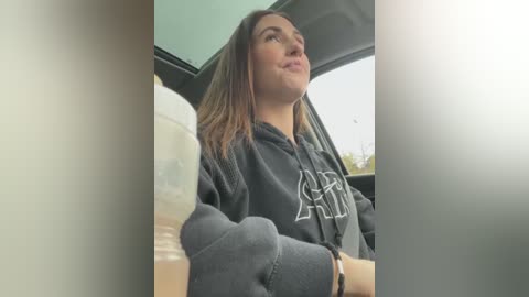 Media: Video of a young woman with shoulder-length brown hair, wearing a dark hoodie, driving a car, holding a white cup, smiling, and gazing upward.