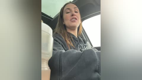 Media: Video of a young woman in a car, wearing a dark hoodie, holding a cup, with a relaxed expression, blurry background.