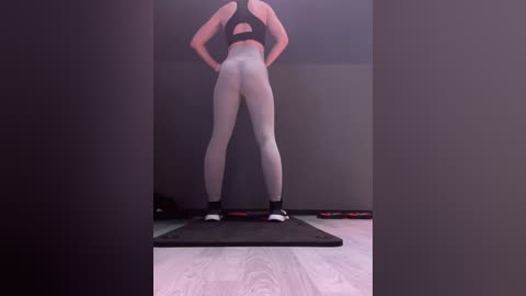 Media: Video of a fit woman with fair skin, wearing a black sports bra and white leggings, standing with arms raised, on a gym mat in a dimly lit room with wooden floor and gray walls.