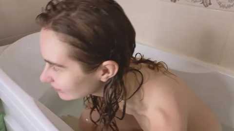 Media: Video of a young, fair-skinned woman with wet, shoulder-length brown hair, topless, sitting in a white bathtub filled with water, looking down.