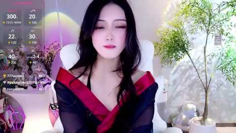 Media: Video of an Asian woman with long black hair, fair skin, and red lipstick, wearing a black kimono with red trim, seated in a modern room with a white chair, potted plants, and digital overlay showing social media stats.