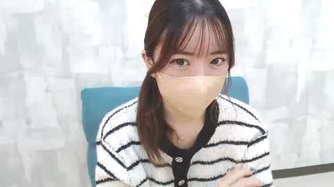 Media: Video of a young Asian woman with fair skin, wearing a white and black striped sweater and a beige face mask, sitting on a blue chair against a white and beige patterned background.