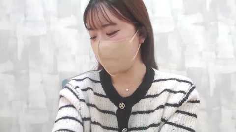 Media: Video of an Asian woman with medium skin tone, shoulder-length dark hair, wearing a beige mask and a black-and-white striped cardigan, against a blurred, patterned white background.