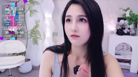 Media: Video of a young East Asian woman with long black hair, fair skin, and a slender physique, wearing a gray tank top, sitting indoors with a modern, minimalist decor featuring white furniture, plants, and a colorful digital game overlay.