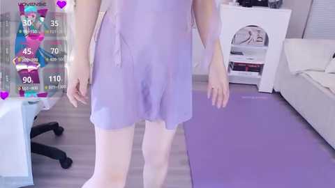 Media: Video of a light-skinned woman in a see-through lavender dress, standing in a modern living room with purple yoga mat, white couch, and colorful digital game stats overlay.