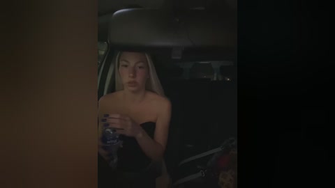 Media: A dimly lit video shows a young, fair-skinned woman with long blonde hair, wearing a strapless black dress, drinking from a blue water bottle inside a dark car.