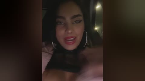 Media: Video of a close-up, dimly-lit, attractive woman with long dark hair, wearing red lipstick and large hoop earrings, smiling seductively, with a blurred background.