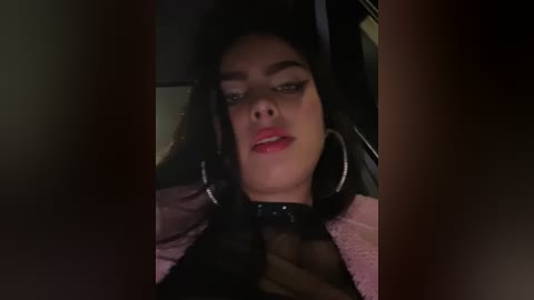 Media: Video of a woman with medium skin tone and dark hair, wearing black sheer top, red lipstick, and large hoop earrings, in a dimly lit room.