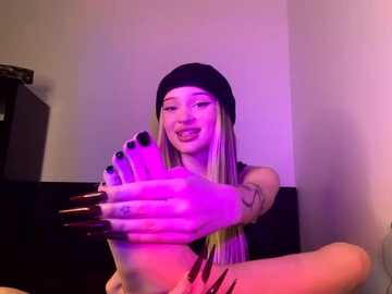 Media: Video of a young woman with long blonde hair, black beanie, and black nail polish, playfully sticking her toes in the air, bathed in purple light.