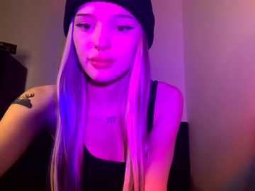 Media: Video of a young Asian woman with long, straight, platinum-blonde hair, wearing a black beanie and black tank top, under purple lighting, showing a tattoo on her upper arm.