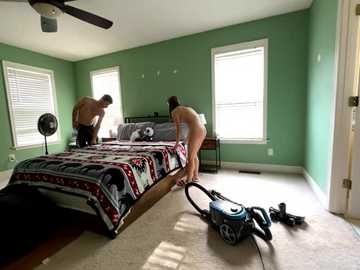 Media: Video of a bedroom with two shirtless men, one vacuuming, another arranging bedding. Green walls, white blinds, and a ceiling fan.