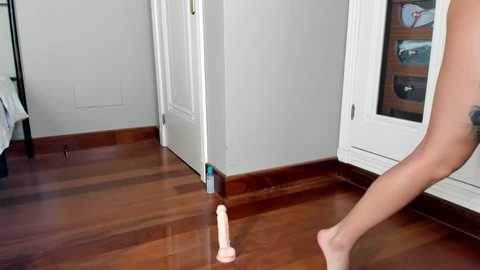 Media: Video of a nude woman standing on a wooden floor, holding a realistic, flesh-colored dildo. Background shows white doors, a window, and a black metal frame.