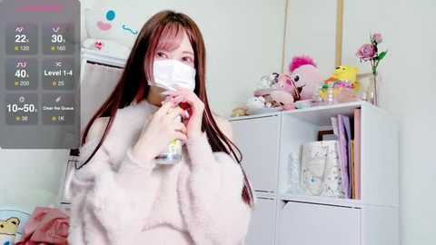 Media: Video of an East Asian woman in a plush, pastel-colored robe, wearing a face mask, standing in a room with white furniture, toys, and a temperature gauge.