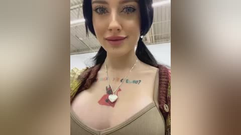 Media: Video of a fair-skinned woman with dark hair, blue eyes, and a tattoo on her chest, wearing a beige tank top and a maroon cardigan. She's smiling, indoors with a white ceiling.