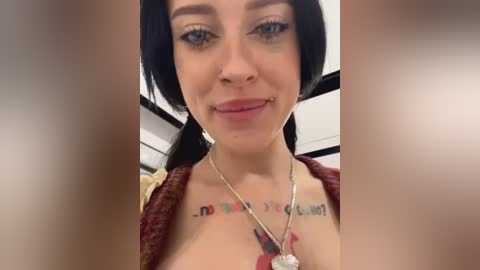 Media: Video of a smiling woman with fair skin, blue eyes, and black hair in pigtails, wearing a maroon top. Her chest features a colorful tattoo. Background shows a blurred, indoor setting with white walls and black ceiling tiles.