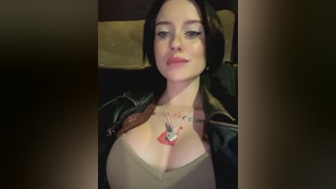 Media: Video of a fair-skinned woman with short black hair, wearing a low-cut beige top, black leather jacket, and a heart necklace. She's seated in a dimly lit car.