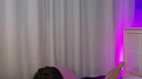 Media: Video of a person with dark hair lying on a bed, reaching out to touch a light switch. The background features white curtains and a purple glow from a light source.