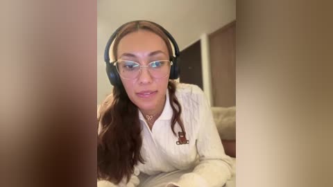 Media: Video of a woman with long brown hair, wearing glasses, a white shirt, and black headphones, lying on a bed, in a dimly lit room.