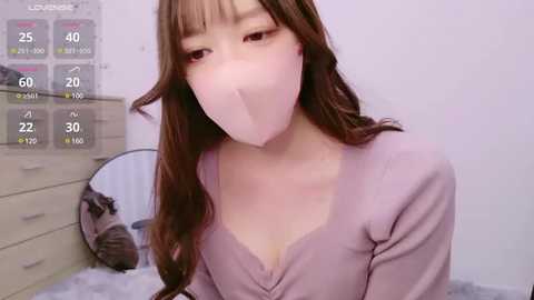 Media: Video of a young Asian woman with long brown hair, wearing a pink face mask, a light purple cardigan, and a white blanket in a bedroom.