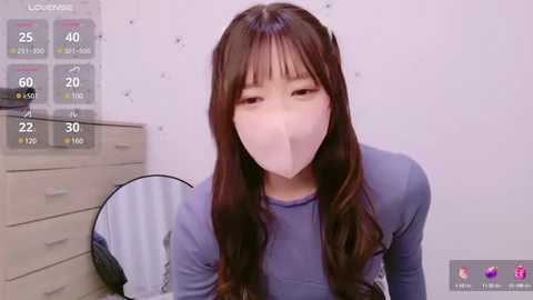 Media: A video of an East Asian woman with long, wavy brown hair and a light complexion, wearing a white face mask, a grey long-sleeve top, and a black skirt. The background includes a white wall, a grey dresser, and a black fan.