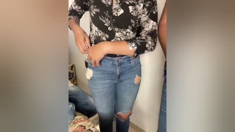 Media: Video of a person wearing a black floral blouse and distressed blue jeans, standing against a plain beige wall in a room with a patterned couch.