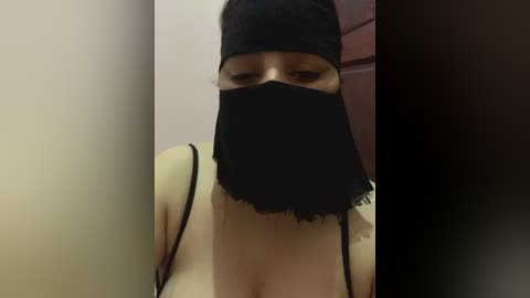 Media: Video of a woman with light skin, wearing a black face mask and black bra, standing against a beige wall.